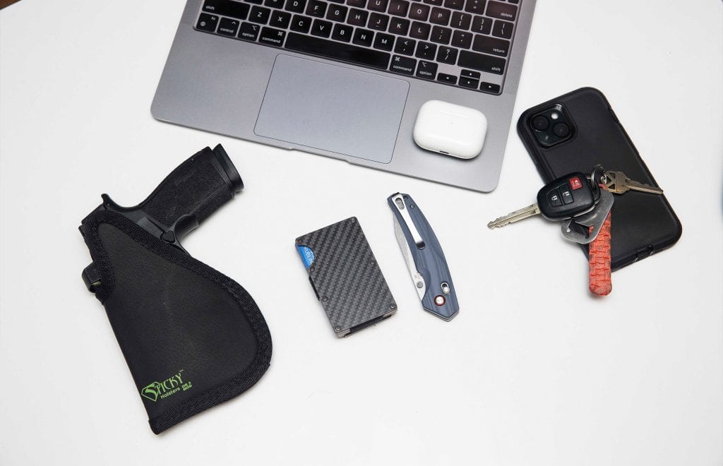 op-down view of a Sticky Holster EDC setup with handgun, carbon fiber wallet, knife, keys, and a laptop.