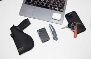 op-down view of a Sticky Holster EDC setup with handgun, carbon fiber wallet, knife, keys, and a laptop.